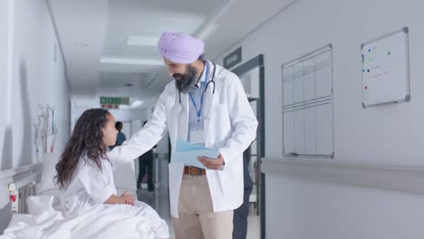 diverse sikh male doctor in turban and child patient talking in corridor at hospital, in slow motion