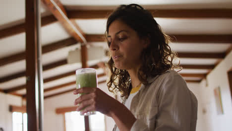 feliz mujer de raza mixta bebiendo bebida de salud de pie en la cabaña sala de estar sonriendo