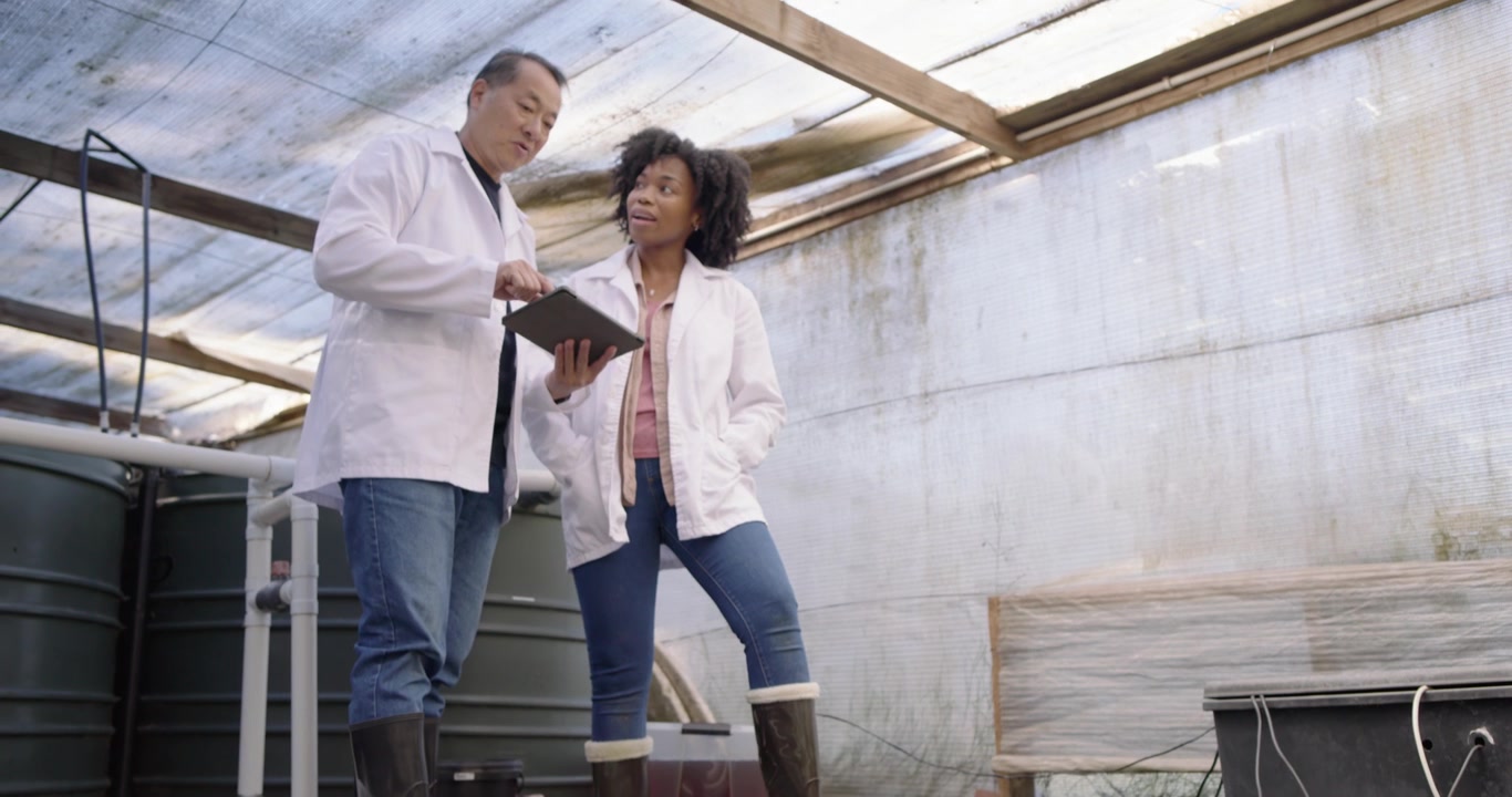 Diverse team in lab coats inspecting tank on deck while PVC pipes pouring water consulting tablet