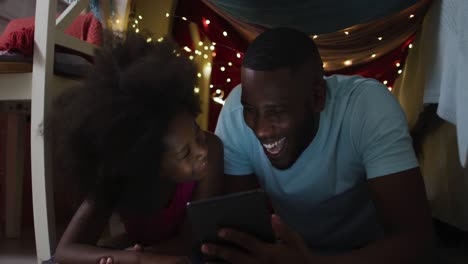 african american daughter and her father watching tablet in blanket tent with fairy lights