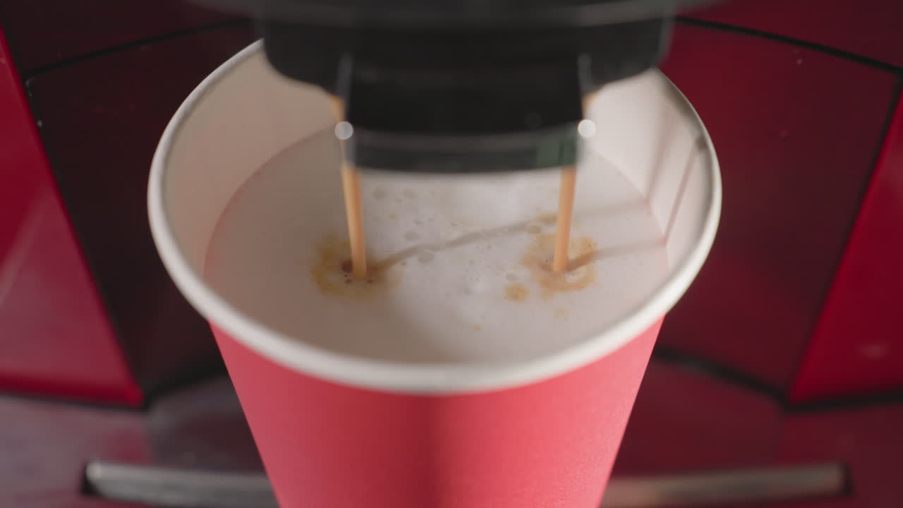 Close-up shot of espresso coffee machine dispensing two streams of hot espresso into red paper cup filled with milk foam, creating a rich, layered coffee drink