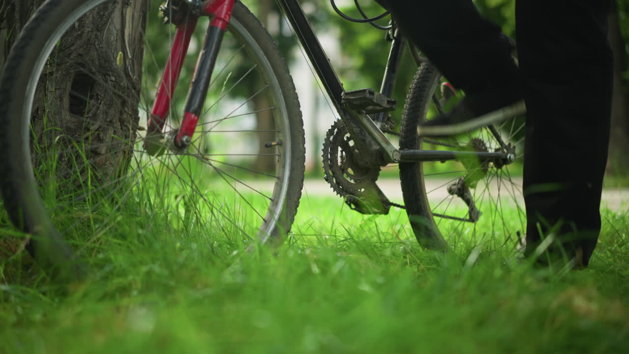주차된 자전거의 페달에 발을 는 개인의 클로즈업, 잔디에 있는 나무에 기대어, 페달 위치를 확인하는 동안 발목을 만지는 사람, 배경에 흐릿한 초록색