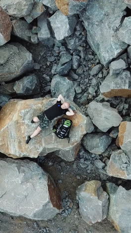 bald man lays on a large stone. tourist with a backpack in shorts and t-shirt is resting. aerial view vertical video