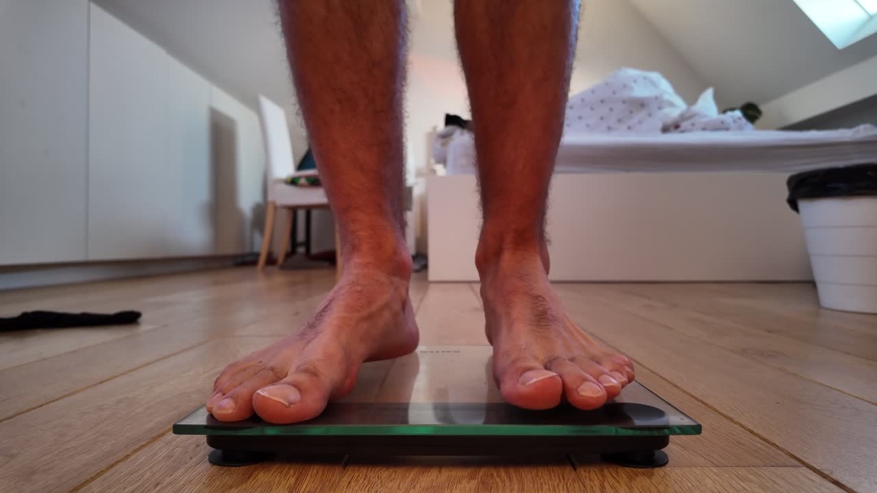 Barefoot man walks toward glass scale on bedroom floor