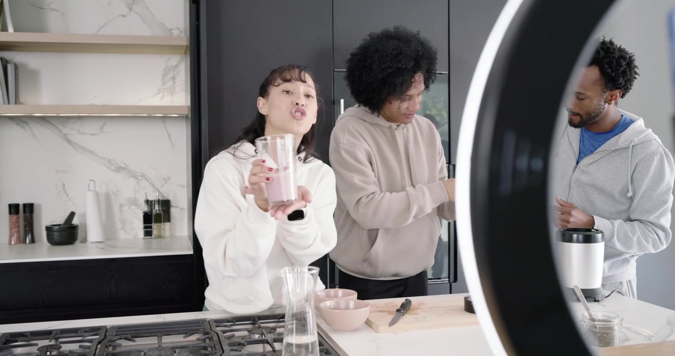 Co-conduttori diversi che accendono il telefono e la ring light, filmando una dimostrazione di frullato con frullatore sul bancone della cucina.
