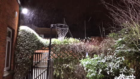 snow falls past a netball hoop in an english cottage garden lit up at night under a flood light