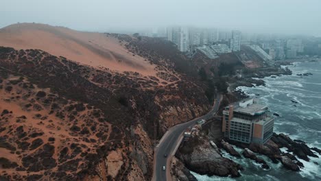 Aerial-truck-right-of-orange-sand-dunes-and-coastal-road-in-rugged-hillside-near-sea-waves-and-Concon-buildings-on-a-foggy-day,-Chile