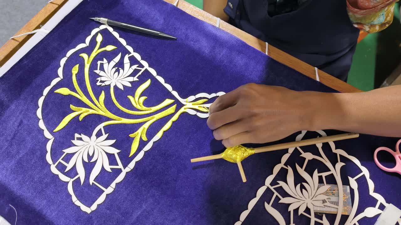 Seorang perajin terampil menampilkan sulaman Tekat Melayu tradisional. Ia dengan cermat menyulam benang emas di atas kain beludru mewah. Kerajinan kuno yang langka ini mewakili bentuk seni warisan yang semakin menghilang.