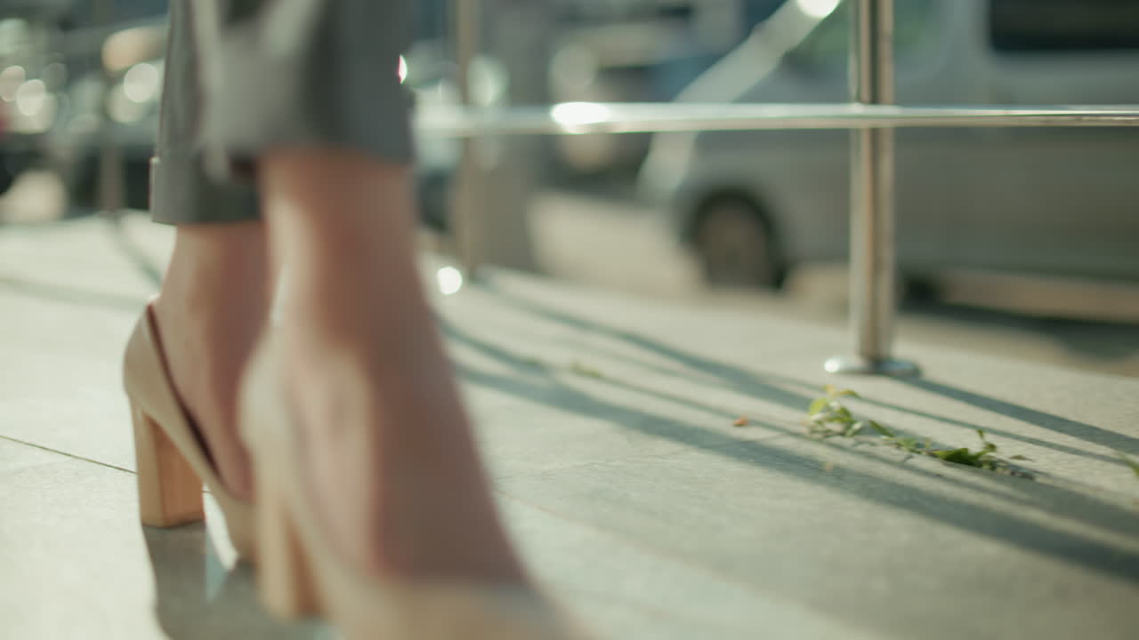 Close up leg view of woman in heels walking gracefully beside iron railing with parked cars in background, sunlight casting soft shadows on pavement as she moves confidently