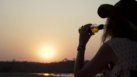 Sonnenuntergangaufnahme-Einer-Frau,-Die-Ein-Getränk-Trinkt,-Während-Sie-Eine-Angelrute-An-Einem-Friedlichen-Fluss-Hält