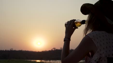 sunset shot of a woman drinking a beverage while holding a fishing rod on a peaceful river