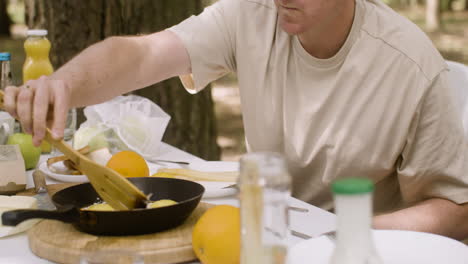 happy man moving fried eggs in a pan with a wooden fork while having breakfast with his family at the camping in the forest