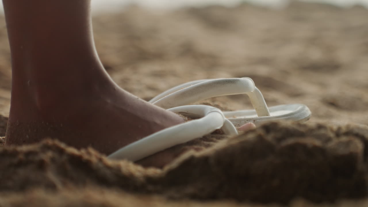 Sandalo appoggiato sulla sabbia calda, primo piano lento di un piede nudo che scivola in un sandalo con cinturino bianco sulla battigia, granelli dorati che aderiscono alla pelle e al cinturino, luce soffusa del tramonto, umore contemplativo, vacanziere in pausa