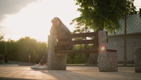 lady seated at the edge of a park bench during golden hour, resting her head in a contemplative manner, as sunlight shines brightly, with a blurred figure walking in the background