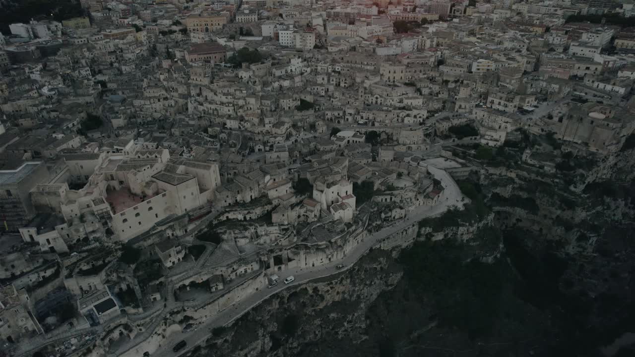 Drone che vola all'indietro con la telecamera che si inclina lentamente verso l'alto, riprendendo l'antica città bianca di Matera al tramonto in 4K.