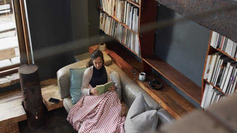 biracial woman lying on sofa under blanket and reading book at home, slow motion