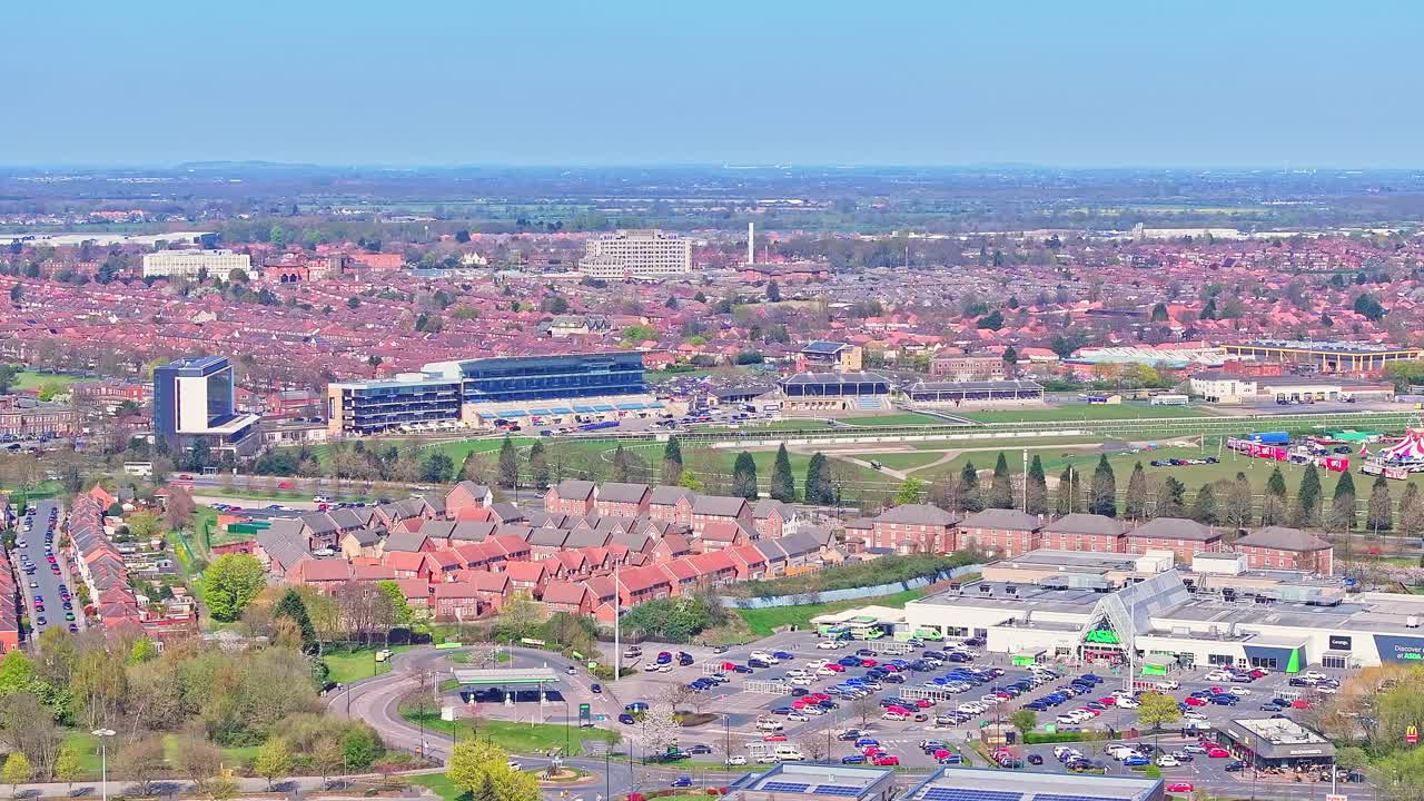 Aerial view during a slow drone ascend over Doncaster Racecourse in South Yorkshire, England, revealing the grandstand, suburban housing, commercial areas, and wide landscape under a clear blue sky