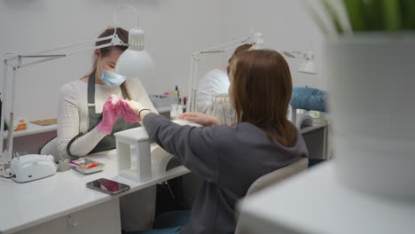 nail technician wearing pink gloves and face mask focused on customer nails under bright white lamp while customer with light brown hair sits with phone on table during manicure session