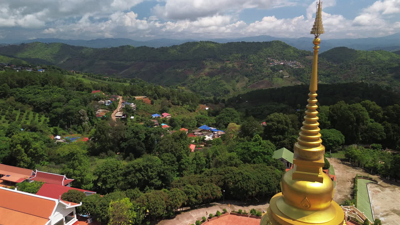 Luftfoto af gylden bakketop-stupa ved Wat Phra That Santitham, Mae Salong, Chiang Rai, omgivet af grønne områder ved middagstid, flyv forbi tempel til landsby