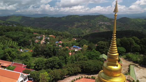 aerial of golden hilltop stupa at wat phra that santitham, mae salong, chiang rai, surrounded by greenery at midday, fly past temple to village
