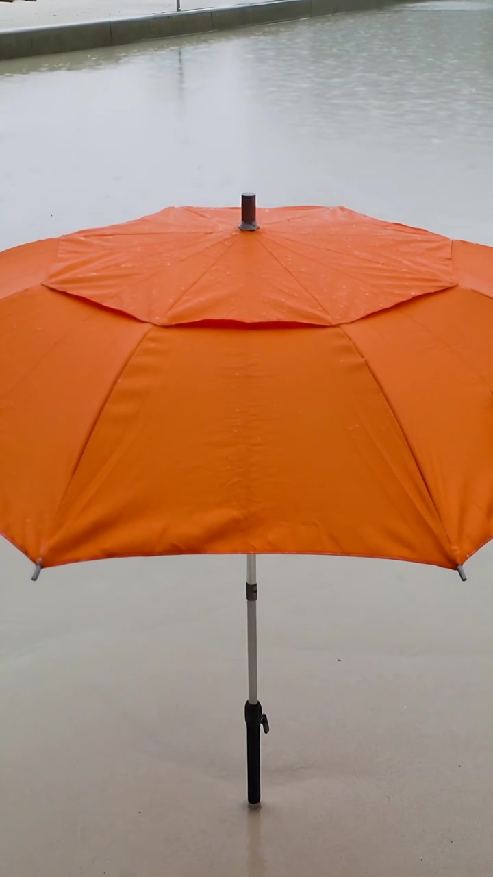 Video verticale: Ombrello arancione in piedi che ripara un patio bagnato mentre la pioggia crea increspature sull'acqua.