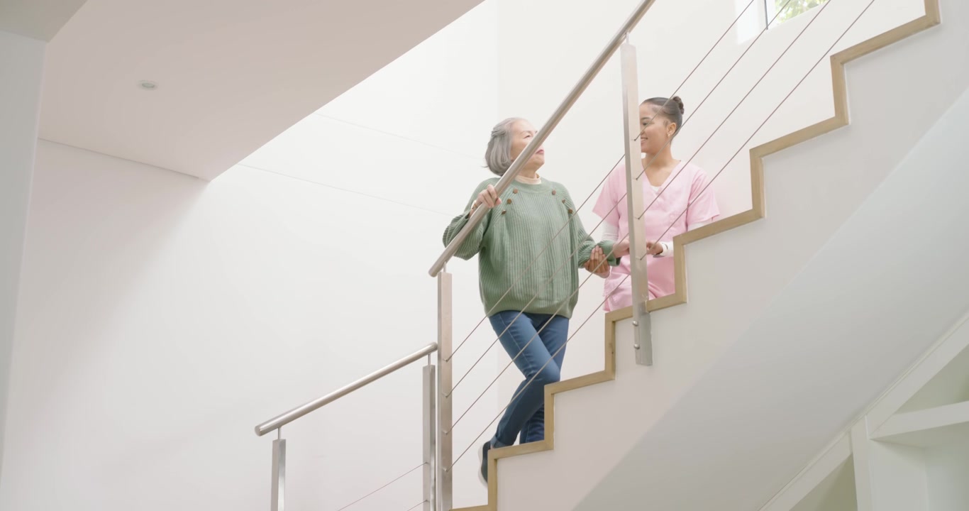 Entrando sulle scale di casa, donna anziana di etnia diversa e assistente sanitario in divisa che salgono, usando il corrimano per sicurezza