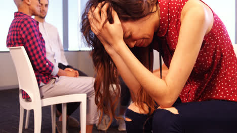 Upset-woman-sitting-on-chair-with-colleagues-discussing-in-background