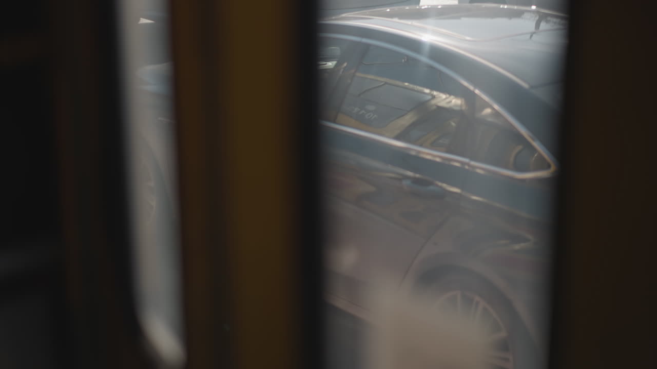 View through moving vehicle window shows car tail rolling along sunlit winter road, soft blur from foggy glass and frames, subtle motion of urban traffic, exhaust haze drifting, reflective asphalt