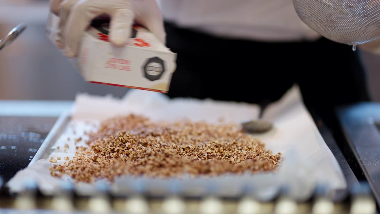 Uno chef che versa un composto di pangrattato e semi in una teglia durante la preparazione del cibo in una cucina trafficata