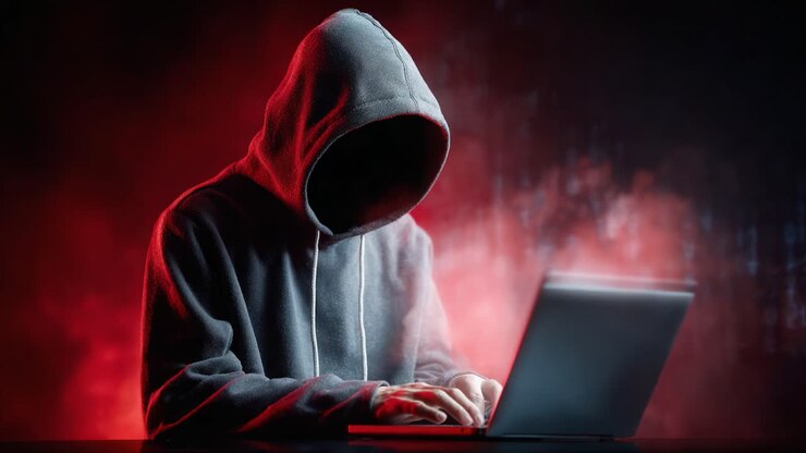 Una figura avvolta nell'ombra con una felpa con cappuccio è intensamente concentrata su un laptop in un ambiente oscuro e suggestivo, illuminato da inquietanti luci rosse e blu, suggerendo temi di mistero e attività informatica.