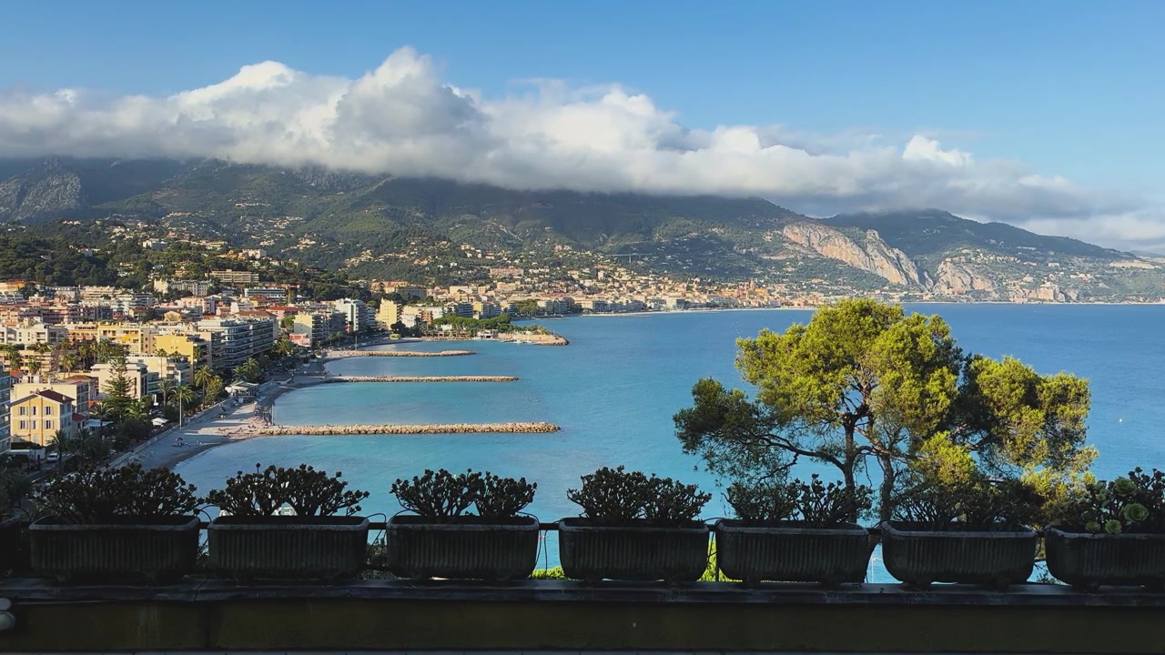 Vista panoramica elevata di Mentone, Francia, lungo la Costa Azzurra. La costa mediterranea, le spiagge, gli edifici sul lungomare e il mare calmo e blu si estendono sotto montagne imponenti parzialmente coperte.