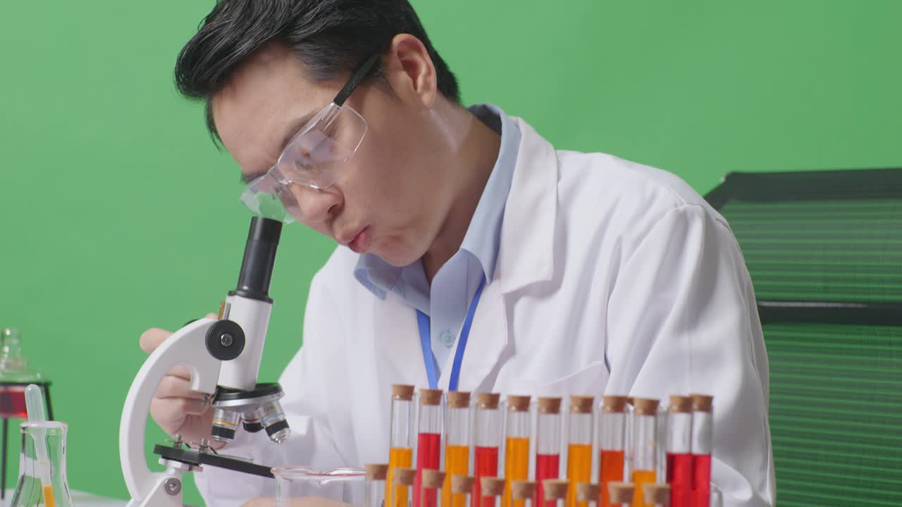 "아시아 남성 과학자가 테이블 위의 현미경을 통해 ""그러면 미소 짓고 와우""라고 말하면서 녹색 스크린 뒷면 실험실에 앉아 있습니다."