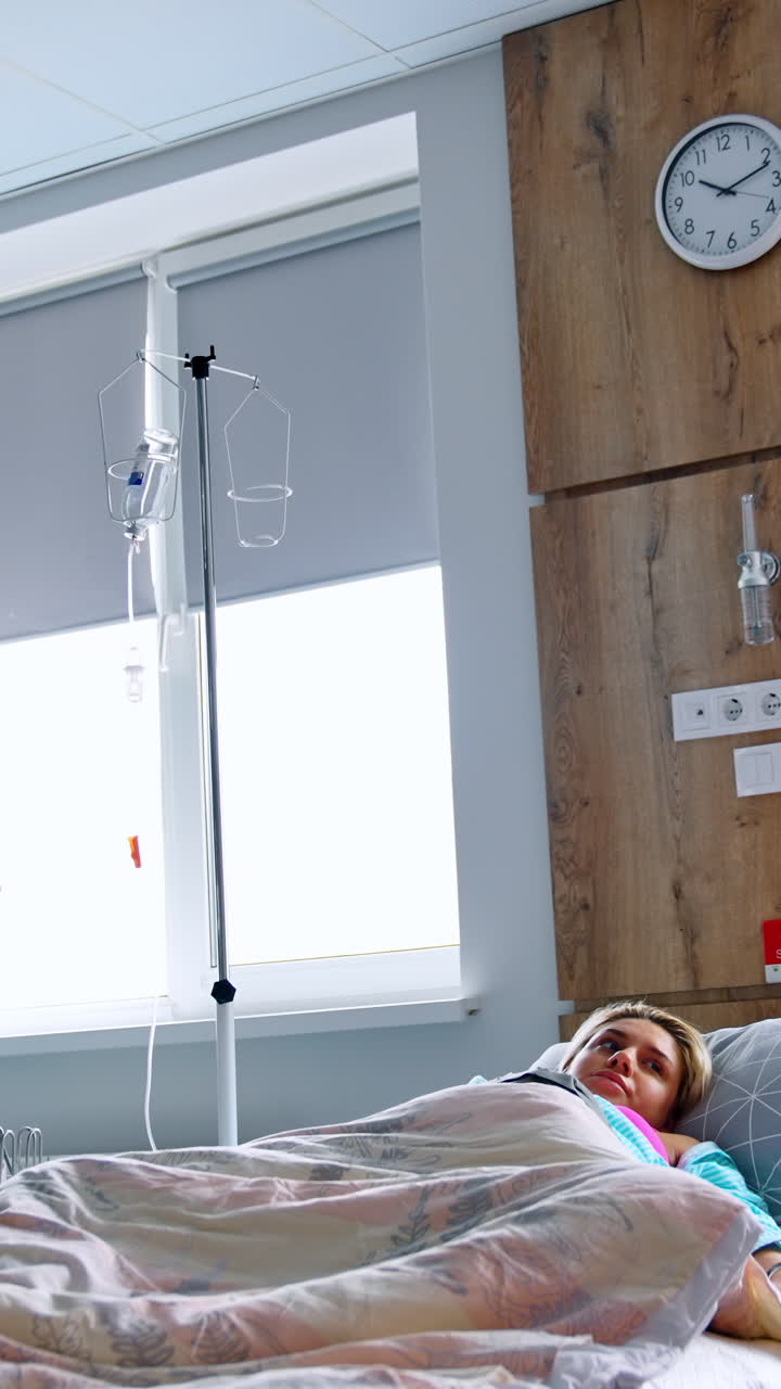 Pregnant woman lies in the hospital bed covered with duvet. Husband sits beside his wife talking and smiling to her. Low angle view. Vertical video