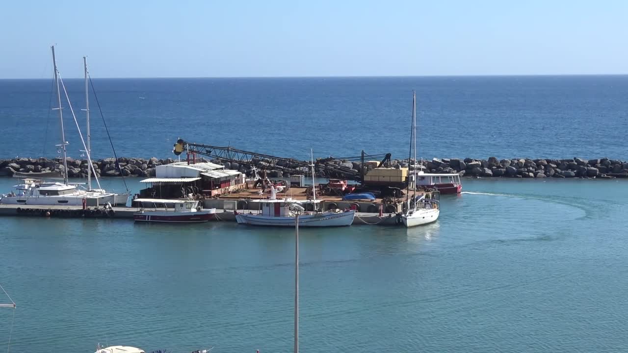 Boats are docked at a shipyard overlooking the sea under a clear blue sky in Greece. Bright sunlight enhances the vibrant colors of the scene.