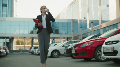 young professional woman confidently walking with coffee and folder through parking lot surrounded by parked cars with modern glass office and residential buildings in background