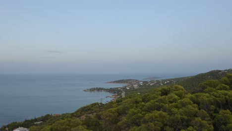 Drone-aerial-view-mountains-covered-with-trees-with-ocean-in-the-background-and-small-rural-village