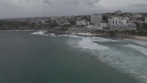 cielo sombrío sobre los suburbios del este - piscina bondi icebergs y club de surf en la playa bondi con surfistas en olas salpicadas en sydney, nsw, australia