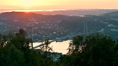 Vista-Desde-Lo-Alto-De-Una-Colina-Que-Domina-El-Puente-Askoy-Al-Atardecer-Con-Luz-Roja-Y-Dorada-En-El-Horizonte-Y-Reflejada-En-El-Agua-A-Lo-Lejos