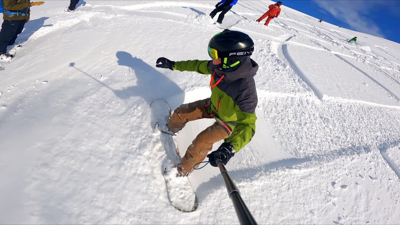 Sportsman taking selfie as he goes down the mountain slope. Lots of people are on the slope at backdrop.
