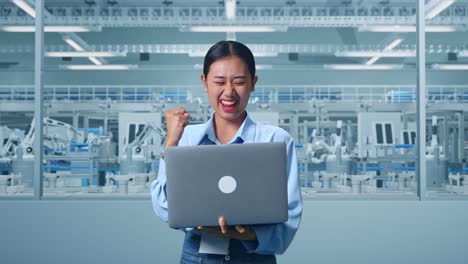 una trabajadora profesional asiática de pie con su portátil en la digitalización de la fábrica para un proyecto innovador, levanta el puño con un objetivo gritante