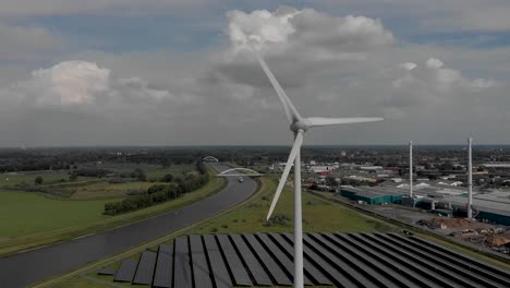 approach and aerial descend showing a field of solar panels beneath a three bladed electricity producing wind turbine against a blue sky with clouds