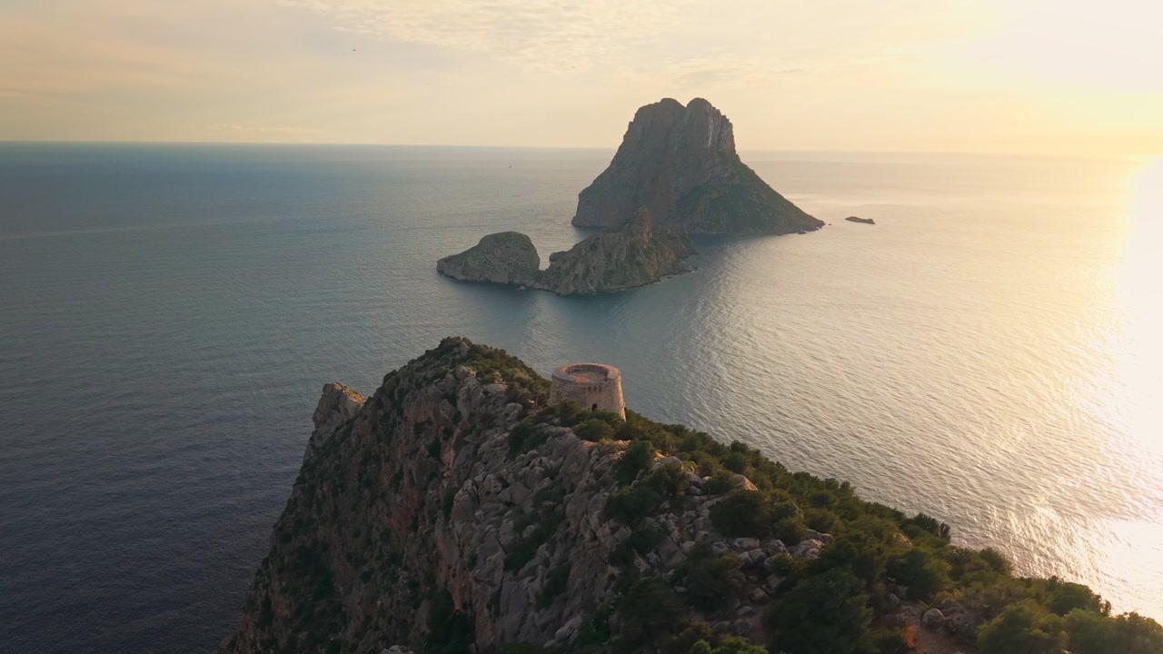 Una vista da drone inquadra una torre in pietra su un promontorio roccioso di fronte a Es Vedrà, Ibiza, Spagna, con la luce del tramonto che si riflette sul Mar Mediterraneo