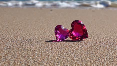 Two heart shaped gemstones, one slightly larger than the other, rest on a sandy beach, their facets catching the sunlight as gentle waves wash ashore