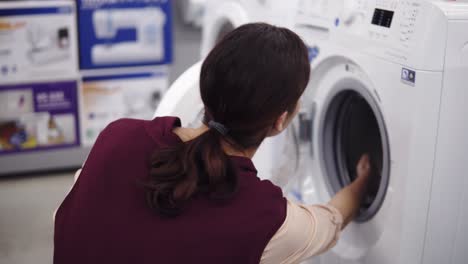 Young-brunette-girl-opens-a-washing-mashine-door,-spins-the-cylinder-inside-and-pulls-out-detergent-tray.-Costumer-looking-for-household-equipment.-Home-appliance.