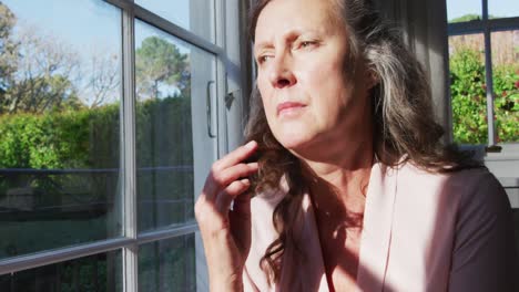 Thoughtful-senior-caucasian-woman-sitting-in-sunny-living-room-looking-out-of-window