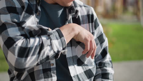 close up of person in black and white checkered shirt pulling medicine from chest pocket while seated outdoors under bright sunlight with blurred green grassy background and distant wooden structure