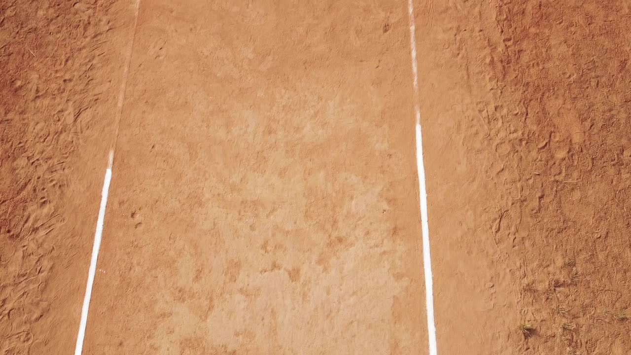Detaljeret dronekamerabevægelse i fugleperspektiv langs en rød ler-cricketbane, der fremhæver hvide markeringslinjer og et sæt røde stumps