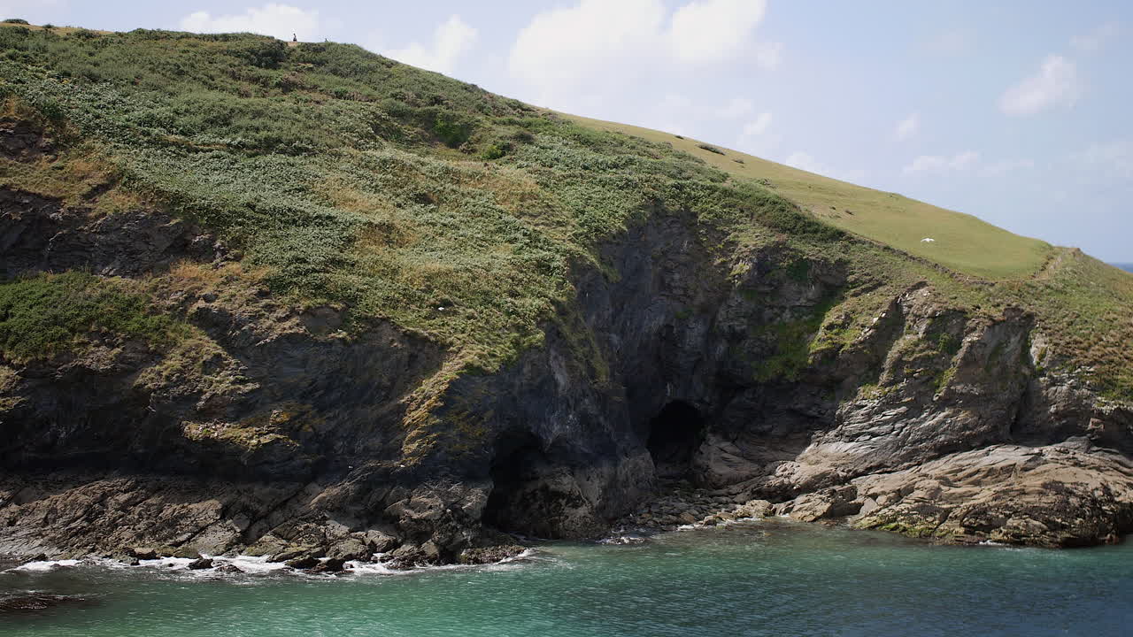 Cliffs with sea caves and grassy slopes are rising above turquoise waters along the Cornwall coast, near the area used for filming Portwenn in Port Isaac