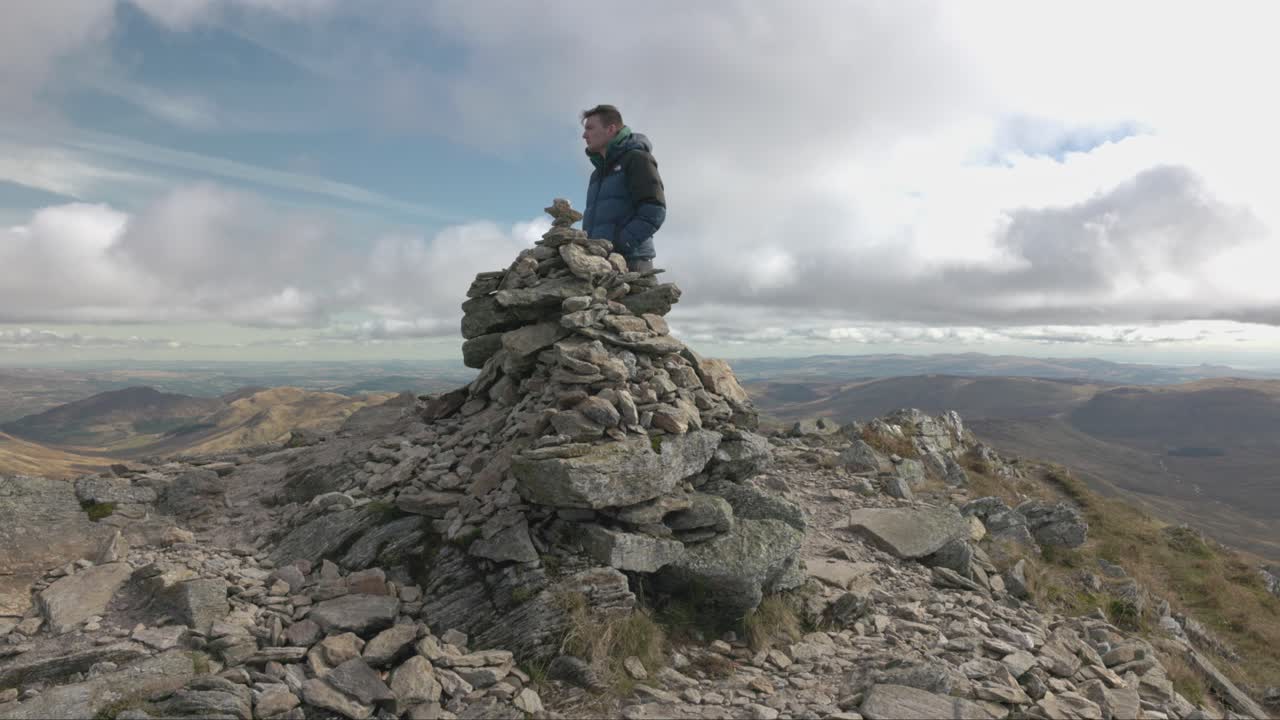 Wide angle shot of a hiker walking up to a stone stack on the summit of Ben Vorlich