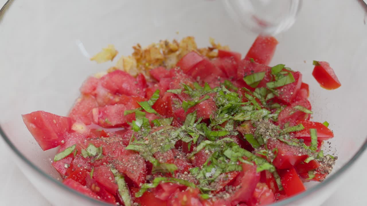 Top-down close-up of chopped tomatoes and basil being seasoned and mixed with spoons in a white bowl